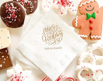 May include: A Christmas-themed display with white napkins printed with gold "Merry Christmas" text and "The Richards". The napkins are surrounded by chocolate-covered sweets, gingerbread biscuits, candy canes, and snowflake-shaped sweets.