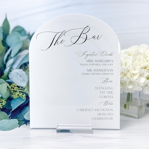 May include: White acrylic sign with a rounded top, featuring the text "The Bar" in elegant calligraphy. The sign lists signature drinks, beer, and wine options, including "Mrs. Margarita", "Mr. Manhattan", "Yuengling", "Fat Tire", "Corona", "Cabernet Sauvignon", "Moscato", and "Chardonnay".