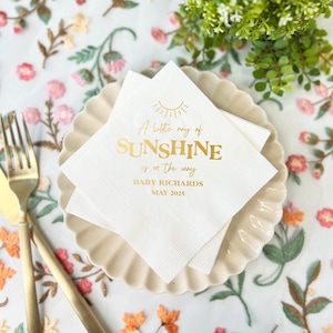 May include: White paper napkins with gold text that reads "A little ray of SUNSHINE is on the way" and "BABY RICHARDS MAY 2025". The napkins are stacked on a beige scalloped plate, with gold silverware on the side.