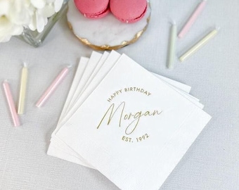 May include: White paper napkins with gold text that reads "Happy Birthday Morgan Est. 1992". The image also shows pink macarons, candles, and flowers. The napkins are stacked on a light-colored surface.