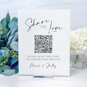 May include: A white sign with the text "Share the Love" and a QR code. Below the code, it reads "Please scan this code to upload your photos!" The names "Mark & Shelly" are at the bottom. The sign is held by a clear acrylic base.