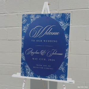May include: Navy blue wedding welcome sign with white floral accents and text. The sign reads "Welcome to our wedding" and includes the names Angelina & Julianne, and the date May 15th, 2026. The sign also says "Thank you so much for coming!"