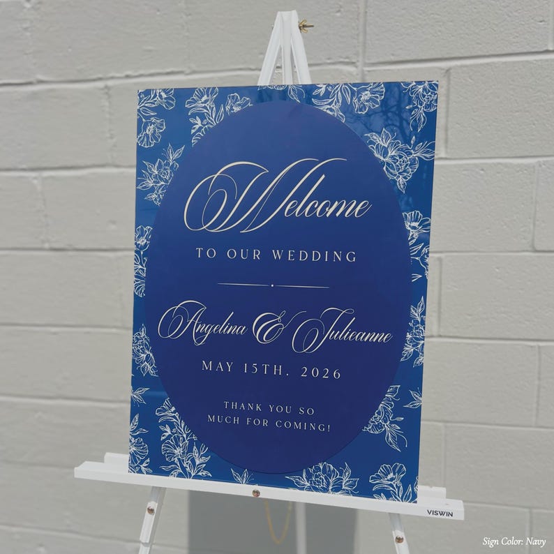 May include: Navy blue wedding welcome sign with white floral accents and text. The sign reads "Welcome to our wedding" and includes the names Angelina & Julianne, and the date May 15th, 2026. The sign also says "Thank you so much for coming!"