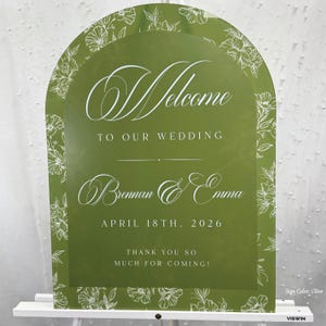 May include: Olive green wedding welcome sign with white floral accents and text. The sign reads "Welcome to our Wedding" followed by names and the date "April 18th, 2026". The bottom of the sign says "Thank you so much for coming!"
