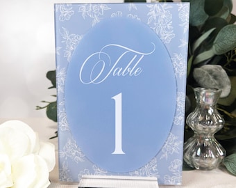 May include: A light blue table number card with the word "Table" and the number "1" in white script. The card has a floral pattern and sits in a clear acrylic stand. Wedding decor.