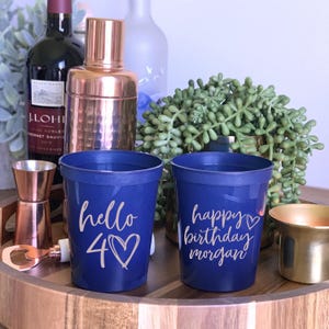 May include: Two navy blue plastic cups with white lettering. One cup says "hello 40" with a heart, the other says "happy birthday morgan". The cups are on a wooden tray with bar accessories and a bottle of wine.