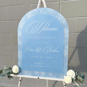 May include: A light blue wedding welcome sign with white floral accents and text. The sign reads "Welcome to our wedding" with the names Olivia & Nathan and the date June 20, 2026. The sign is on a white easel.