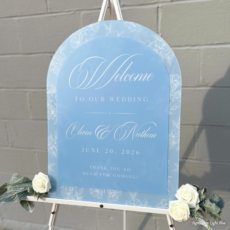 May include: A light blue wedding welcome sign with white floral accents and text. The sign reads "Welcome to our wedding" with the names Olivia & Nathan and the date June 20, 2026. The sign is on a white easel.