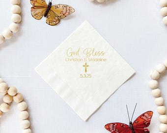 May include: Ivory napkin with gold text reading "God Bless", names, a cross, and the date 5.3.25. Two decorative butterflies and wooden beads surround the napkin on a white surface.