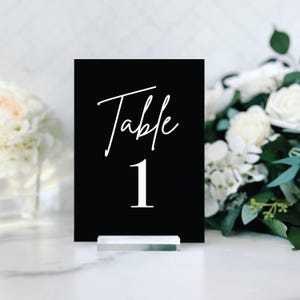 May include: Black table number card with white text that reads "Table 1" on a clear acrylic stand.
