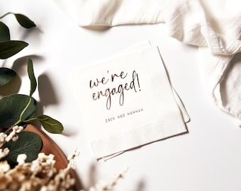 May include: White napkins printed with the words "we're engaged!" and the names "Zach and Hannah" in brown. The napkins are on a white surface, with a white cloth and foliage in the background.