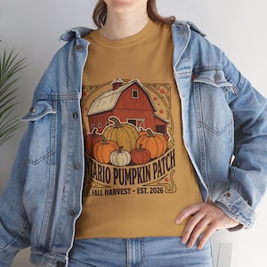 May include: A mustard yellow t-shirt featuring a graphic of a red barn and various pumpkins. The text reads ONTARIO PUMPKIN PATCH FALL HARVEST - EST. 2026. The shirt is worn under a light blue denim jacket with metal buttons and long sleeves.