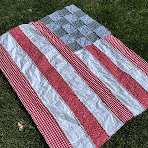 May include: A handmade quilt in the shape of the American flag, displayed on green grass. The quilt features red and white stripes, a blue square with white stars, and red and white gingham trim.