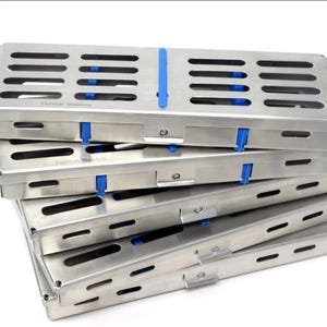 Puede incluir: Una pila de cinco casetes de esterilización rectangulares de acero inoxidable con tapas ranuradas e inserciones de silicona azul. Cada caja de metal cuenta con un mecanismo de cierre con bisagras y el texto German Stainless grabado.
