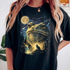 Könnte beinhalten: Schwarzes T-Shirt mit einer detaillierten Illustration einer Kröte, eines Vollmonds und eines Wolfs in einem Wald. Das Gesicht der Kröte steht im Mittelpunkt, vor einem sternenklaren Nachthimmel. Das Design ist auf einem schwarzen, übergroßen T-Shirt gedruckt.