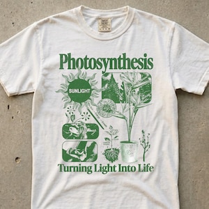 May include: White t-shirt with a green graphic design. The design includes the word "Photosynthesis" above a diagram of plants, the sun, and the text "Turning Light Into Life". The shirt has a crew neck and short sleeves.