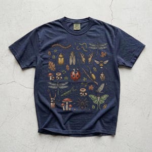 A vintage cottagecore print featuring a detailed collection of insects and woodland foliage. The layout centers on a large ladybug, surrounded by a centipede, dragonflies, a golden beetle, a cicada, a butterfly, a bumblebee, a praying mantis, a stag beetle, a spider, and a green luna moth. Interspersed among the detailed bugs are various red and brown mushrooms, autumn oak leaves, twigs, and tiny mystical stars. The artwork utilizes muted earthy tones for a classic nature aesthetic.