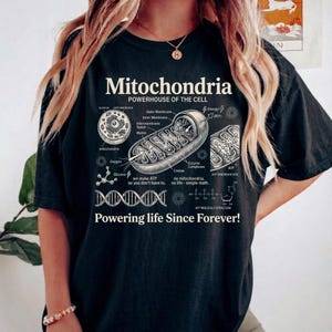 A cream-colored educational biology print titled "Mitochondria" and "POWERHOUSE OF THE CELL" in a bold serif font. The layout features detailed vintage engraving-style diagrams including a cross-section of a mitochondrion with labels like Cristae and Matrix, a cell diagram, a DNA strand, and an ATP molecular structure. Slogans read "we make ATP so you don't have to" and "Powering life Since Forever!" at the bottom. The entire design has a clean, high-contrast textbook aesthetic.