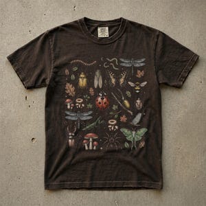 A vintage cottagecore print featuring a detailed collection of insects and woodland foliage. The layout centers on a large ladybug, surrounded by a centipede, dragonflies, a golden beetle, a cicada, a butterfly, a bumblebee, a praying mantis, a stag beetle, a spider, and a green luna moth. Interspersed among the detailed bugs are various red and brown mushrooms, autumn oak leaves, twigs, and tiny mystical stars. The artwork utilizes muted earthy tones for a classic nature aesthetic.