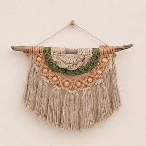 May include: A macrame wall hanging suspended from a piece of driftwood. The design features concentric semicircular layers in sage green, cream, and terracotta, accented with light wooden beads and finished with long, beige fringe tassels.