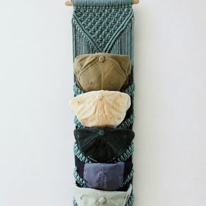 May include: A vertical wall-mounted macrame organizer in a muted teal color, featuring five integrated pockets. Each pocket holds a baseball cap in varying shades of olive, cream, dark green, navy blue, and light sage, suspended from a wooden dowel.