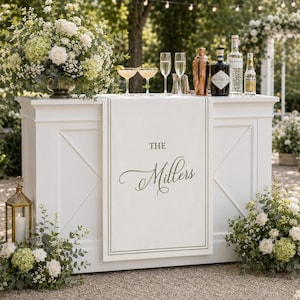 May include: A white wooden bar counter featuring a custom fabric runner with a dark green border and the text "THE Millers" printed in a script font. The bar is decorated with floral arrangements, glassware, and various beverage bottles.