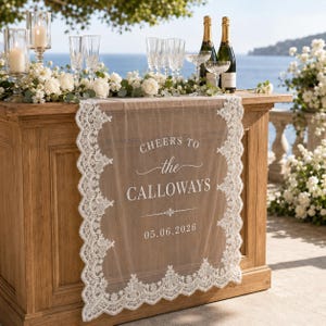 May include: A sheer white lace table runner draped over a wooden bar, featuring the printed text "CHEERS TO the CALLOWAYS" and the date "05.06.2026". The runner has an intricate scalloped lace border and is decorated with white floral arrangements.