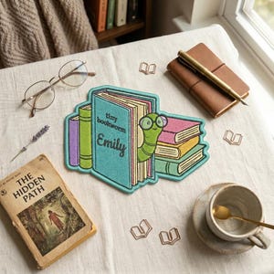 Peut inclure: Un écusson brodé coloré avec un ver de livre sortant d'une pile de livres. Le patch indique "tiny bookworm Emily". On aperçoit également un livre ancien, des lunettes et une tasse de café.