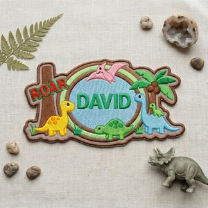 May include: A custom embroidered patch featuring a light blue oval center with the name DAVID in green letters. The patch includes a yellow dinosaur, a green dinosaur, a blue dinosaur, a pink pterodactyl, and a palm tree, all outlined in brown.