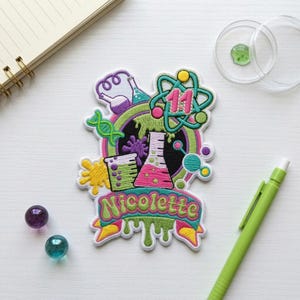 May include: An embroidered patch featuring a science theme with a beaker, flask, DNA strand, and atom symbol. The design includes the number 11 and the name Nicolette in green lettering on a banner. The patch uses bright colors like pink, green, and yellow.