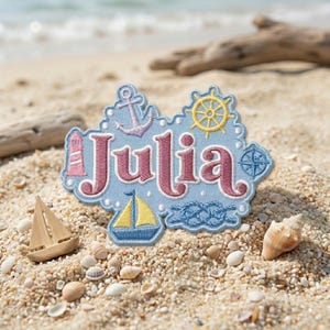 May include: A light blue embroidered patch featuring the name Julia in pink letters. The design includes a pink lighthouse, a purple anchor, a yellow ship wheel, a yellow and blue sailboat, and a blue nautical knot on a light blue background.