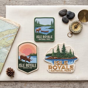 May include: Three embroidered patches featuring Isle Royale National Park. One rectangular patch shows a moose and canoe on a lake. One shield-shaped patch depicts a moose at sunset. One irregular patch shows a forest island with the text Isle Royale.