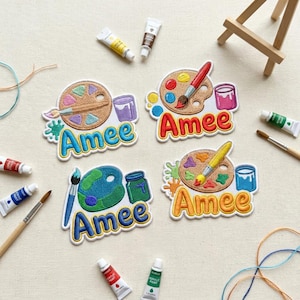 May include: Four embroidered patches featuring a tan or blue artist palette, a paintbrush, a paint jar, and the name Amee in colorful bubble letters. The patches are surrounded by various paint tubes, a wooden easel, and loose colorful threads.