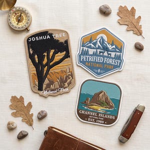 May include: Three embroidered patches featuring Joshua Tree, Petrified Forest, and Channel Islands National Parks. The patches are displayed alongside a brass compass, a folding pocket knife, a brown leather journal, and several small stones and leaves.