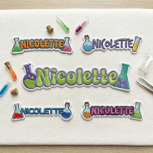 May include: Four embroidered patches featuring the name Nicolette in various fonts and colors, each accompanied by laboratory glassware icons like beakers and flasks filled with colorful liquids, arranged on a white fabric surface.