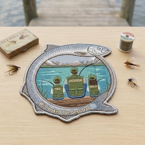 May include: A circular embroidered patch shaped like a fish, featuring a scene of three people fishing on a dock with mountains in the background. The text reads "JACKSON," "PAUL," "ASHLEY," and "We hooked the best Dad" in white embroidery.