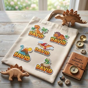 May include: A beige canvas tote bag featuring five embroidered dinosaur patches, each with the name DAVID in orange letters. The bag is surrounded by two wooden dinosaur figurines, a brass compass, and a brown leather journal labeled David's Adventure Log.