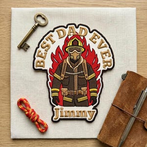 May include: An embroidered patch featuring a firefighter in gear against red flames, with the text "BEST DAD EVER" and "Jimmy" in gold lettering. The patch is accompanied by a metal skeleton key, a red and yellow cord, and a brown leather notebook.