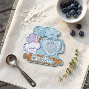May include: An embroidered patch featuring a light blue stand mixer, a purple oven mitt, a chef hat, and a brown rolling pin with the name Emilia stitched in white. The design includes a whisk and spatula, rendered in detailed thread work.