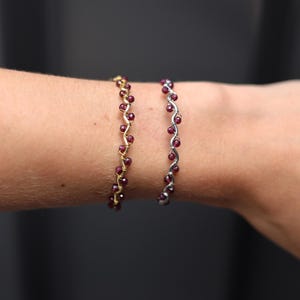 May include: Two bracelets featuring a delicate, wavy metal wire design. One bracelet is crafted from gold-toned wire, while the other is silver-toned. Both are adorned with small, round, deep red garnet beads set along the undulating wire structure.
