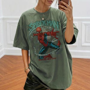 May include: Olive green t-shirt featuring a Spider-Man graphic. The design includes the words "SPIDER-MAN" and "BRAND NEW DAY". The superhero is depicted in his classic red and blue suit, striking a dynamic pose.