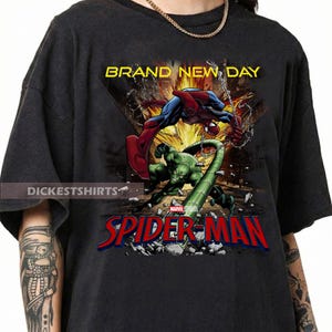 May include: A black short-sleeve t-shirt featuring a graphic print of Spider-Man fighting the Scorpion. The text BRAND NEW DAY appears at the top in yellow, and the red and blue SPIDER-MAN logo is printed at the bottom above a debris-filled scene.