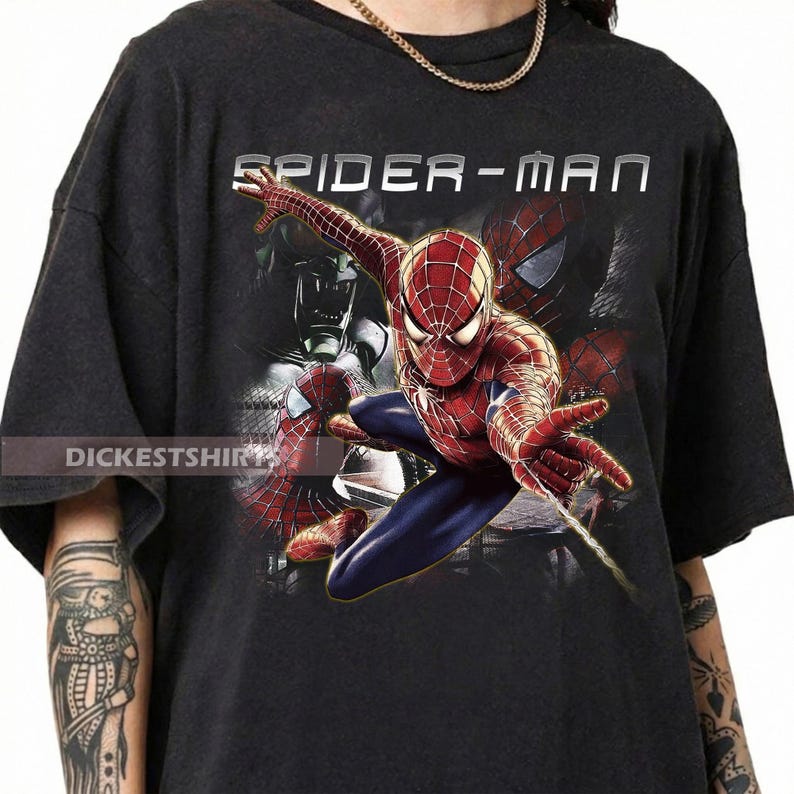 May include: A black short-sleeve t-shirt featuring a graphic print of the character Spider-Man in a dynamic pose with a web-shooting hand. The text SPIDER-MAN is printed in white above the character, and the text DICKESTSHIRT is visible on the left.