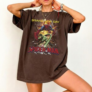 May include: A dark brown short-sleeve t-shirt featuring a graphic print of Spider-Man and the Hulk in a dynamic action scene. The text reads BRAND NEW DAY above the characters and SPIDER-MAN in large red letters at the bottom of the graphic.