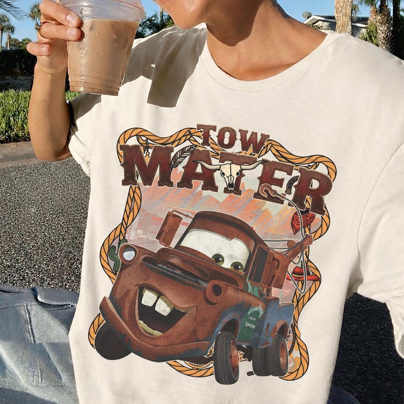 May include: Cream-colored t-shirt featuring a graphic of Tow Mater from the movie Cars. The design includes the text "TOW MATER" in a brown, western-style font, with a cartoon image of the character. The shirt has a classic crew neck.