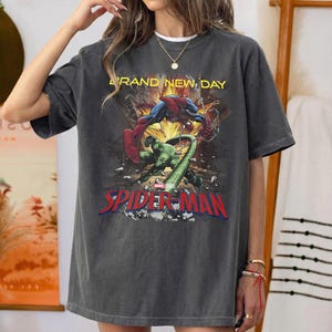 May include: A charcoal gray short-sleeve t-shirt featuring a graphic print of Spider-Man and the Hulk in a dynamic action scene. The text "BRAND NEW DAY" appears in yellow above the characters, with the red "SPIDER-MAN" logo printed below.