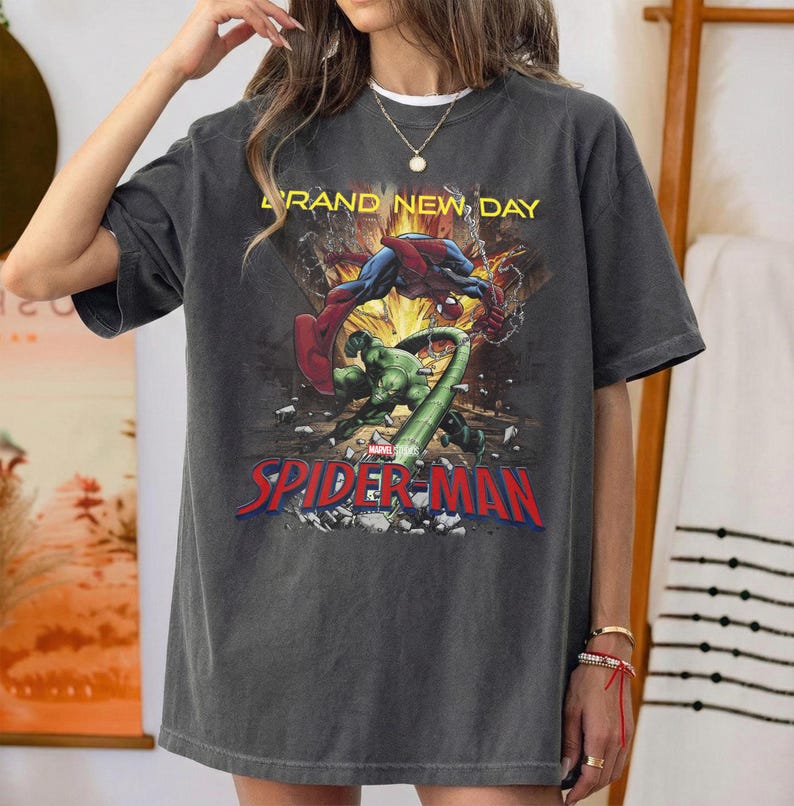May include: A charcoal gray short-sleeve t-shirt featuring a graphic print of Spider-Man and the Hulk in a dynamic action scene. The text "BRAND NEW DAY" appears in yellow above the characters, with the red "SPIDER-MAN" logo printed below.