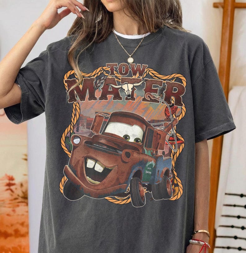May include: Dark gray t-shirt featuring a graphic of Tow Mater from the movie Cars. The design includes the text "TOW MATER" in a western-style font, with a cartoon image of the character. The shirt has a vintage, distressed look.