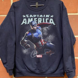 Puede incluir: Sudadera azul marino con Captain America. El dise&ntilde;o incluye el texto "CAPTAIN AMERICA" sobre una imagen de Captain America en acci&oacute;n, una firma y el logotipo de Marvel. La sudadera es de cuello redondo.