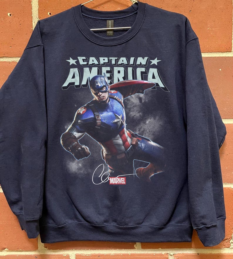 Puede incluir: Sudadera azul marino con Captain America. El dise&ntilde;o incluye el texto "CAPTAIN AMERICA" sobre una imagen de Captain America en acci&oacute;n, una firma y el logotipo de Marvel. La sudadera es de cuello redondo.